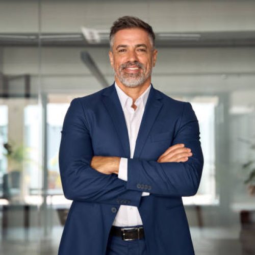 Handsome hispanic senior business man with crossed arms smiling at camera. Indian or latin confident mature good looking middle age leader male businessman on blur office background with copy space.